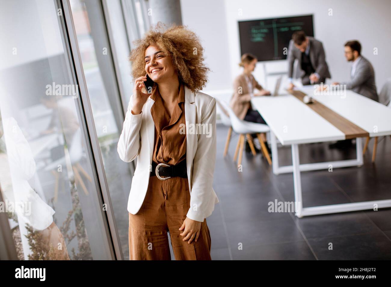 Young curly hair businesswoman using mobile phone in the office with young people works behind her Stock Photo