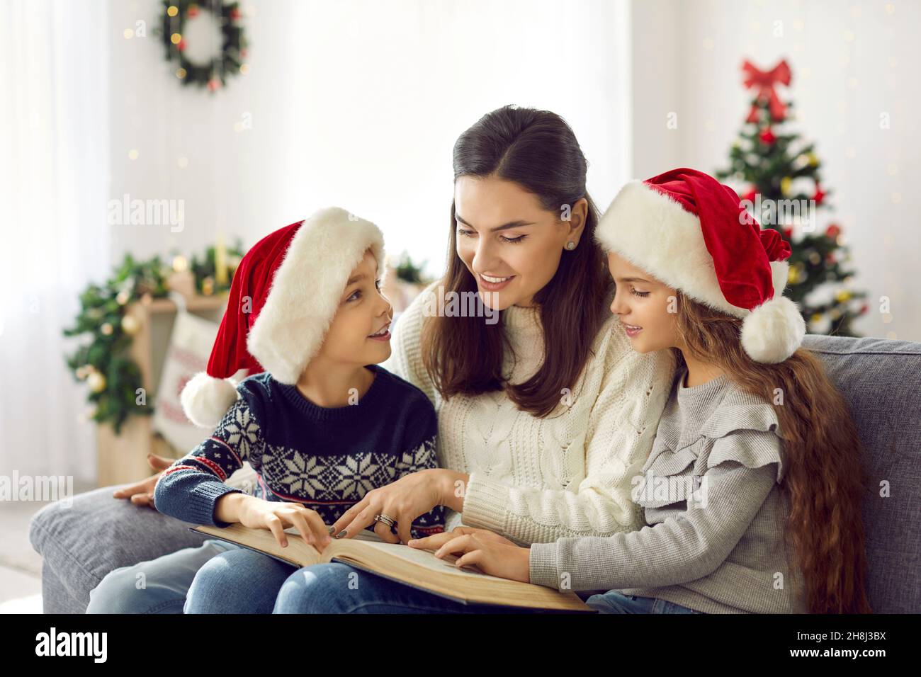 Happy mother and children sitting on couch and reading book on Christmas Eve at home Stock Photo