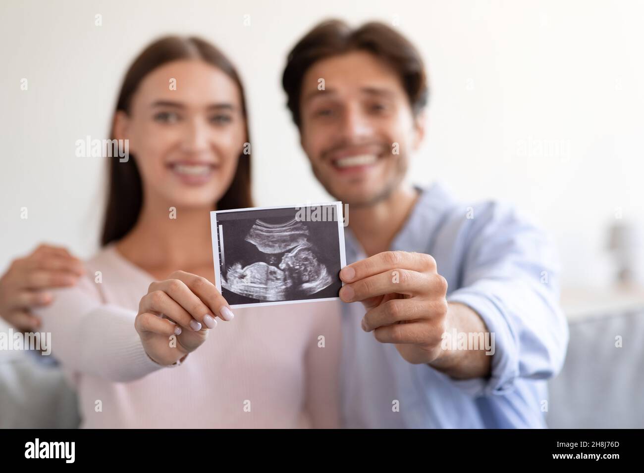 Smiling young attractive european wife and husband hold ultrasound scan picture unborn baby in room Stock Photo
