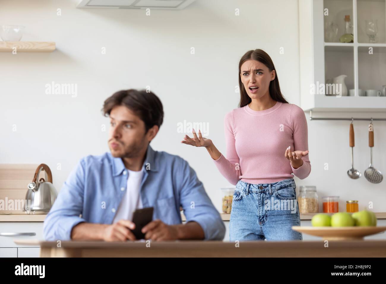 Sad angry upset offended young wife yelling at husband with smartphone on kitchen interior Stock Photo