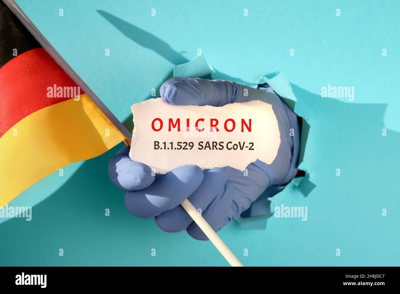 Omicron in Germany, new corona virus variant of concern. Hand in glove from torn paper hole holds German flag and scrap of paper with name of the new Stock Photo