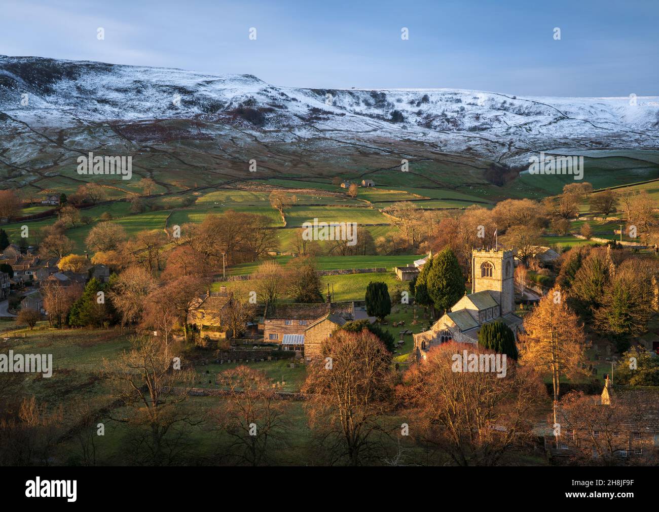 A brief burst of light at sunrise illuminates St. Wilfrid's Church in the pretty village of Burnsall with the hills behind covered in early snowfall. Stock Photo