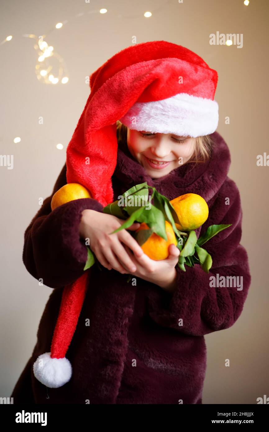 A Caucasian happy girl in a red Santa cap and a fur coat holds a lot of tangerines with leaves. There are lights in the background. Stock Photo