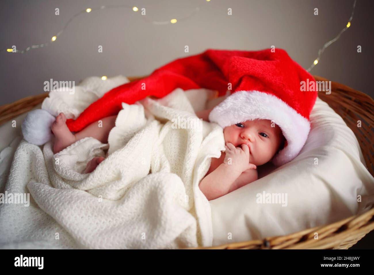 Caucasian newborn baby in Santa's Christmas cap lies in a basket under a white blanket, lights burning against the background. Stock Photo