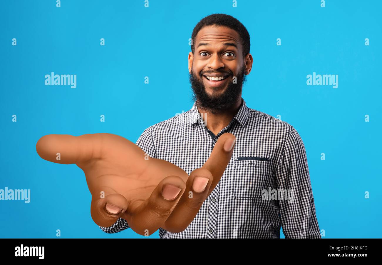Surprised young black man standing with big outstretched hand on blue studio background, mockup for your product Stock Photo