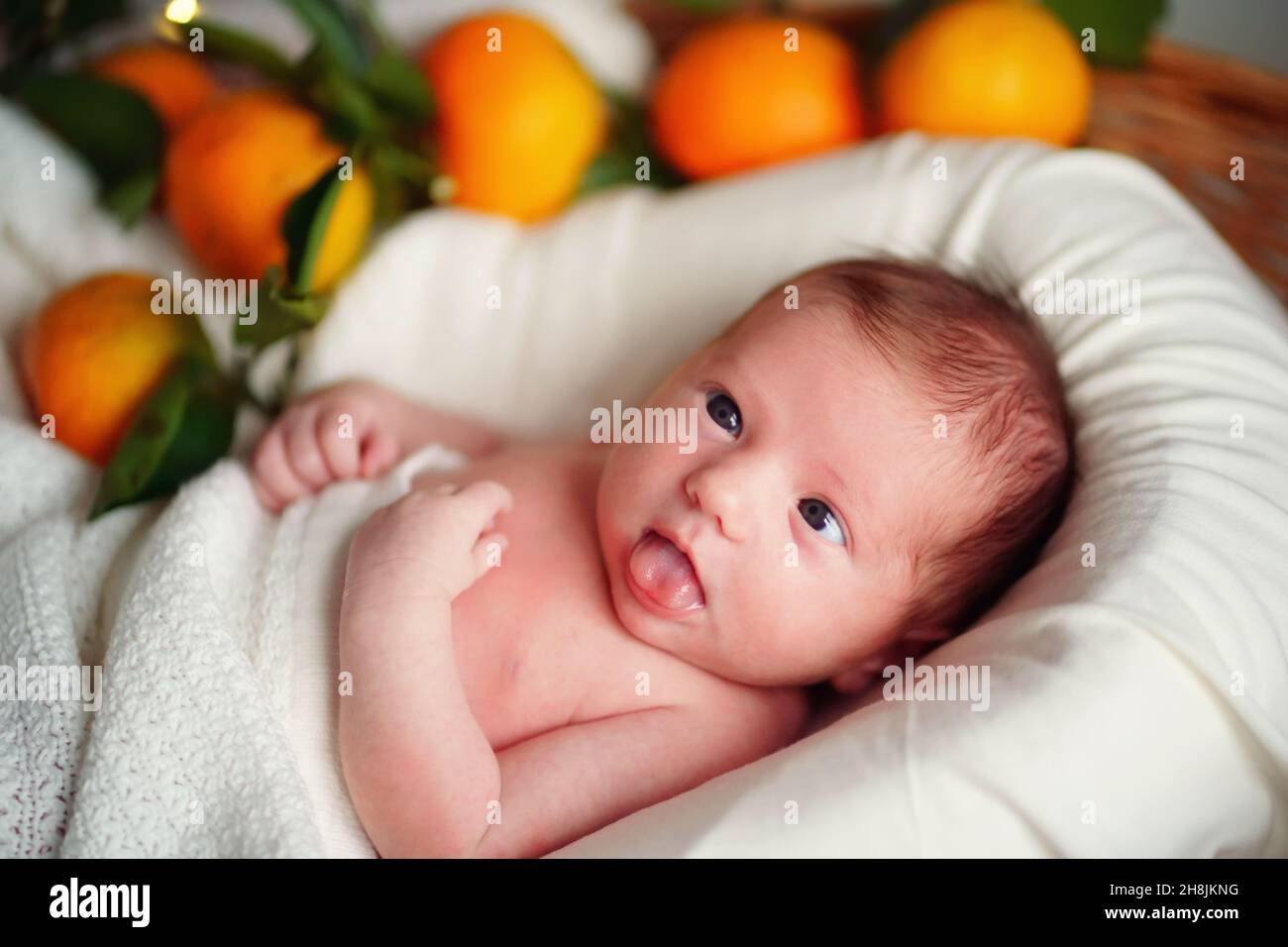 Caucasian newborn cross-eyed baby lies in a basket of tangerines under a white blanket. Stock Photo