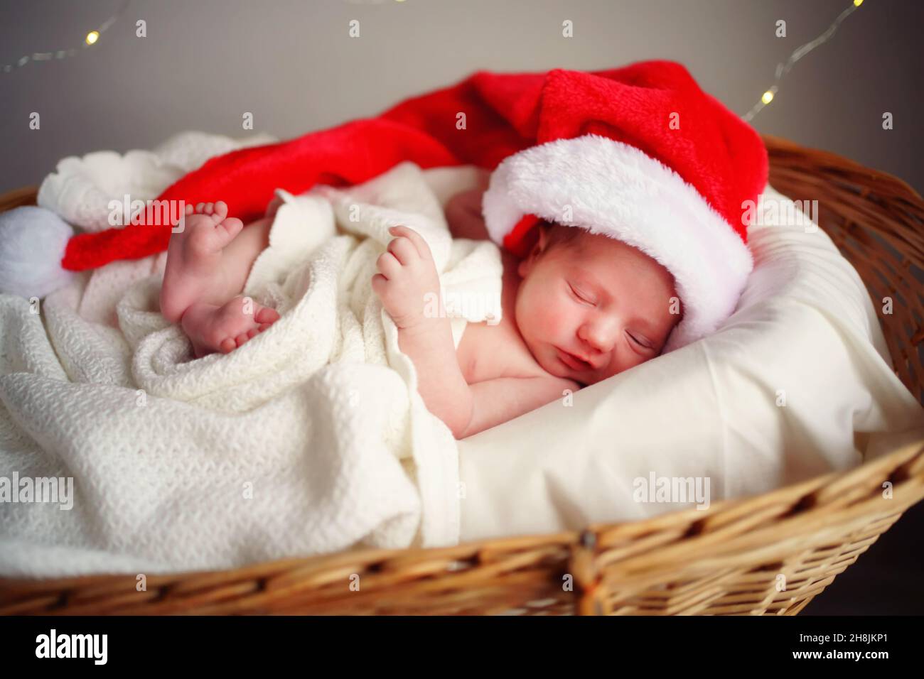 Caucasian sleeping newborn baby in Santa's Christmas cap lies in a basket under a white blanket, lights burning against the background. Stock Photo