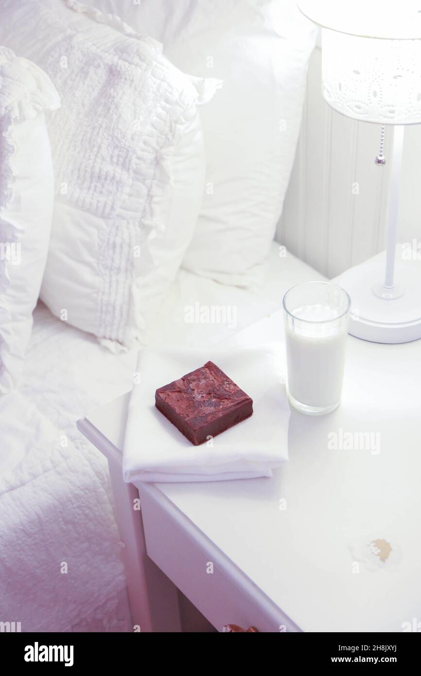 ingle brownie on napkin with glass of milk on night stand next to bed for night time treat.  High key vertical photograph. Stock Photo