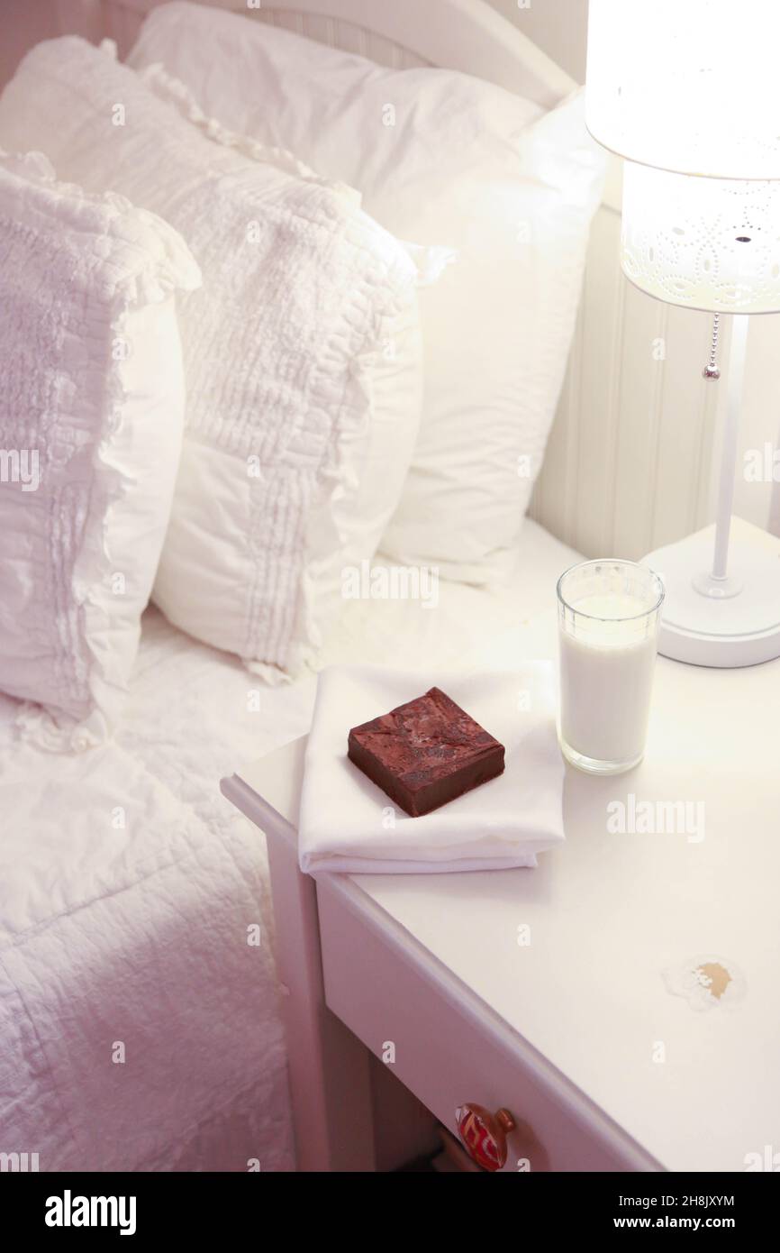 ingle brownie on napkin with glass of milk on night stand next to bed for night time treat.  High key vertical photograph. Stock Photo