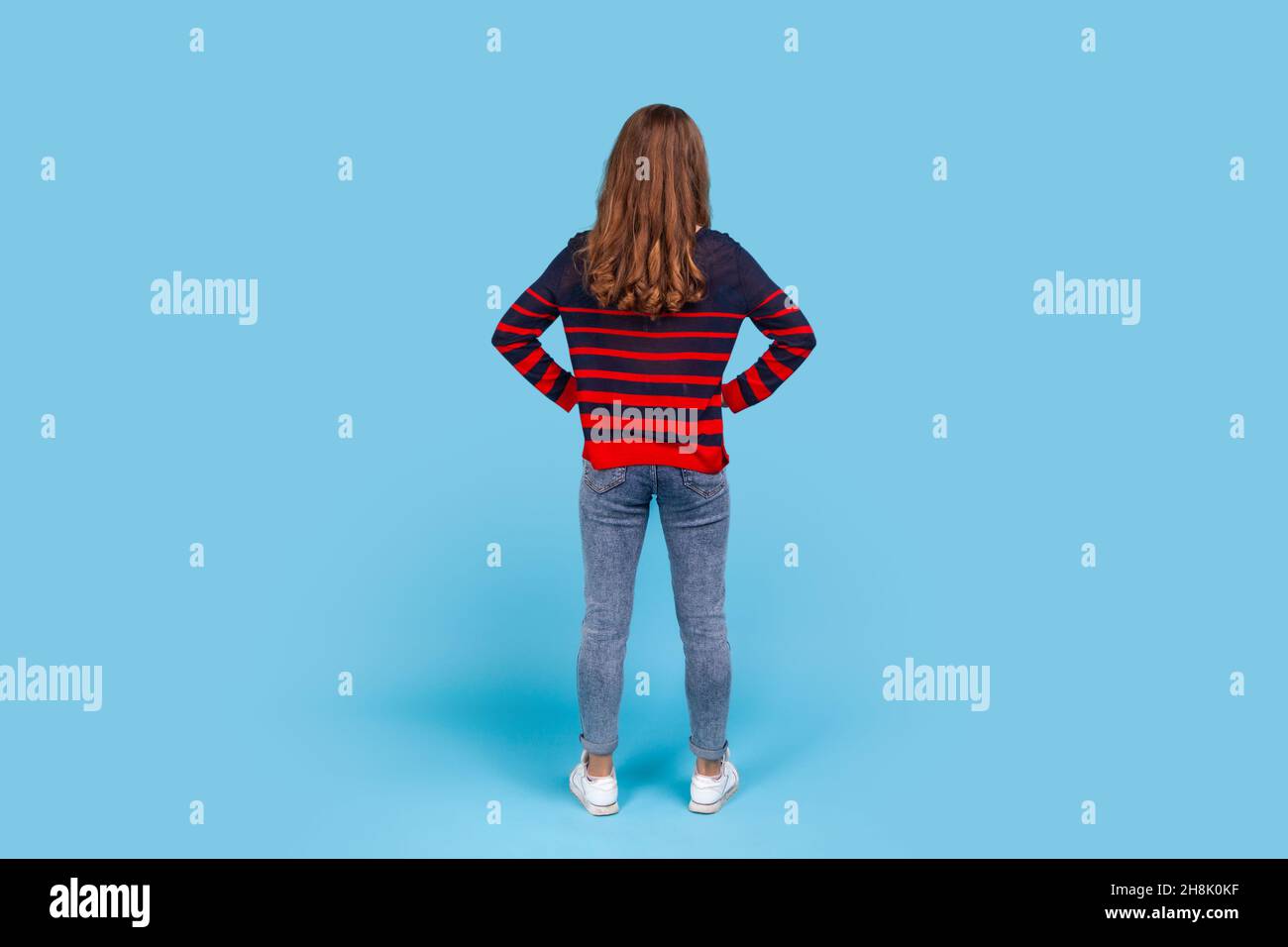 Full length back portrait of woman wearing striped casual style sweater, posing backwards with hands on hips, standing calm unrecognizable, waiting. Indoor studio shot isolated on blue background. Stock Photo