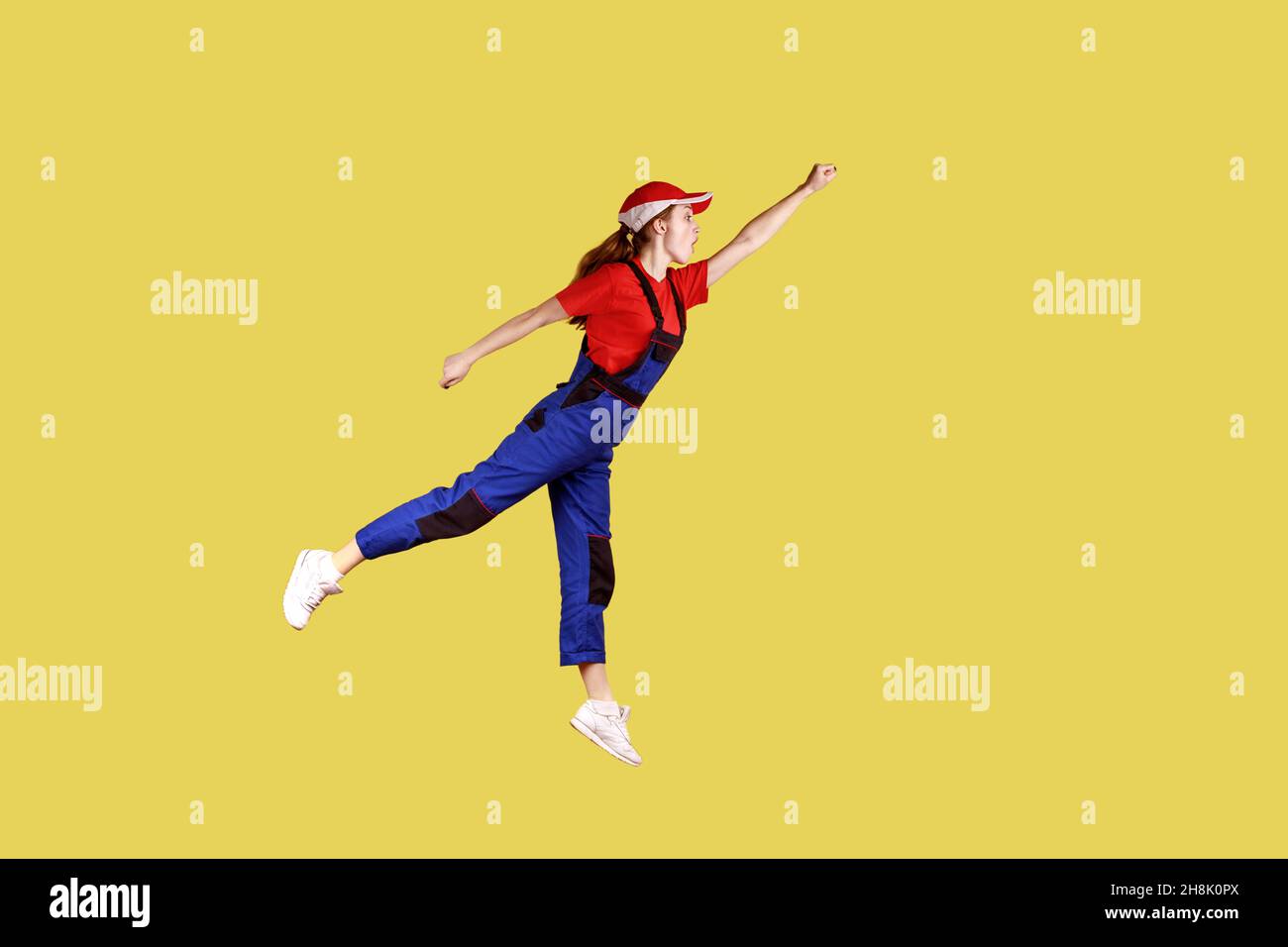 Side view portrait of woman worker flying to do her work like superhero, fast and high quality service, wearing work uniform and red cap. Indoor studio shot isolated on yellow background. Stock Photo
