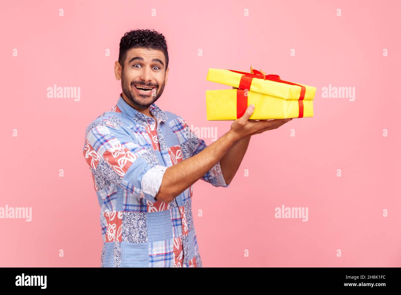 Happy excited man with beard holding in hands half opened yellow wrapped present box, looking at camera with excitement and surprise. Indoor studio shot isolated on pink background. Stock Photo