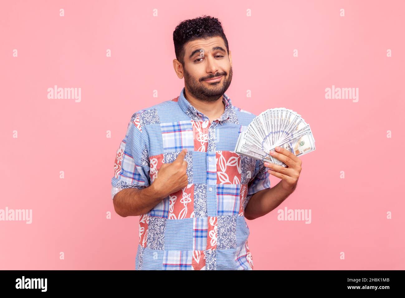 Young adult man with beard and pleasant appearance holding dollars bills and pointing at himself with pride, wearing blue casual style shirt . Indoor studio shot isolated on pink background. Stock Photo