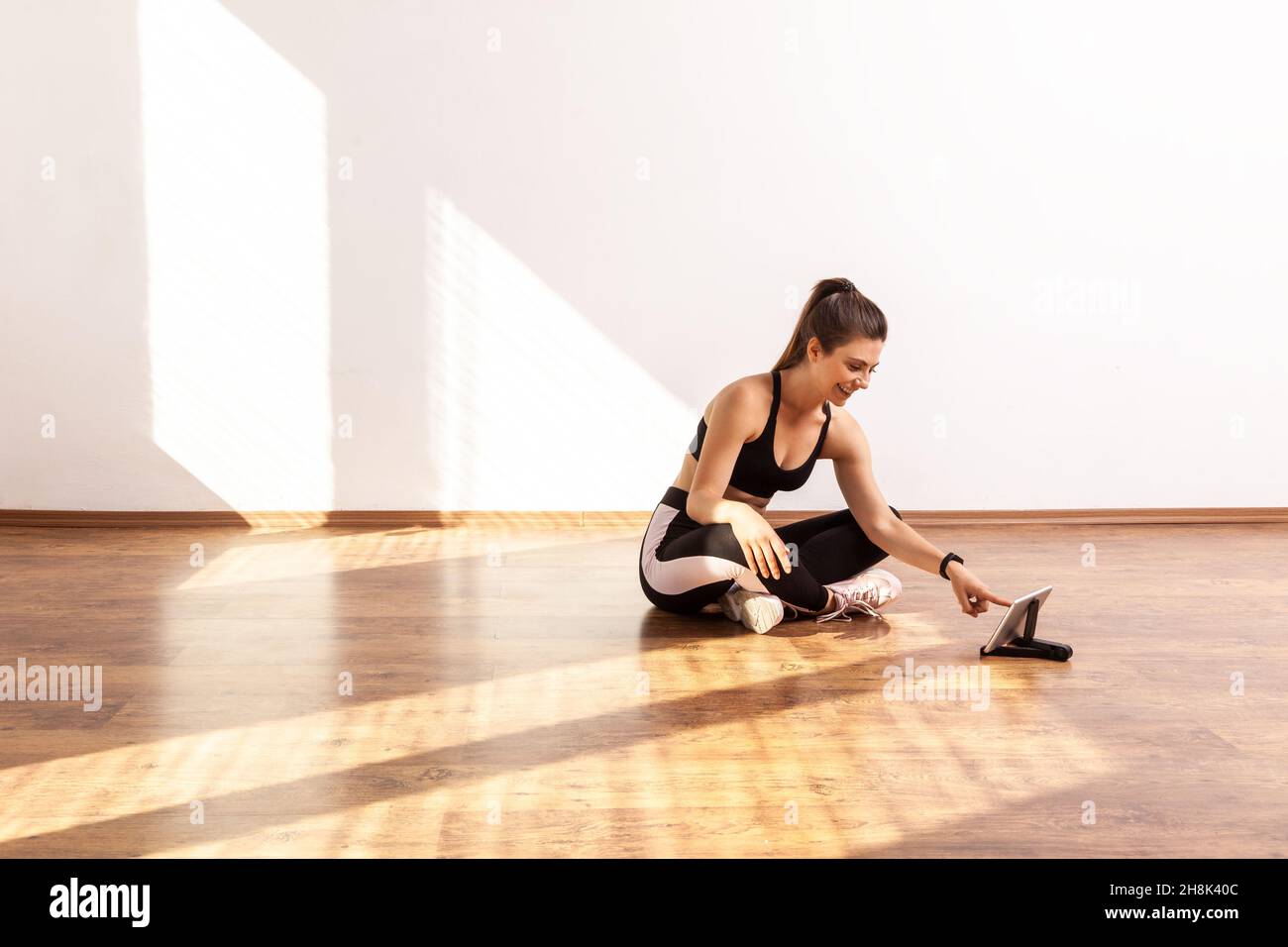 Woman searching online fitness course for beginners in Internet, training video on tablet, wearing black sports top and tights. Full length studio shot illuminated by sunlight from window. Stock Photo