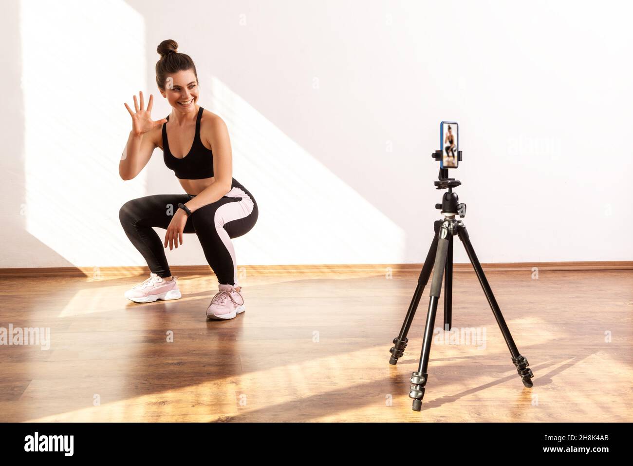 Coach recording video for her fitness vlog, squatting with followers, showing number of remaining, wearing black sports top and tights. Full length studio shot illuminated by sunlight from window. Stock Photo