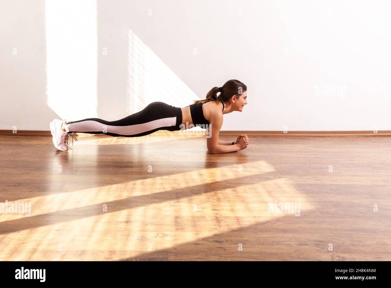 Side view of sportive woman practicing yoga, doing plank exercise on bent hands, training muscles, wearing black sports top and tights. Full length studio shot illuminated by sunlight from window. Stock Photo