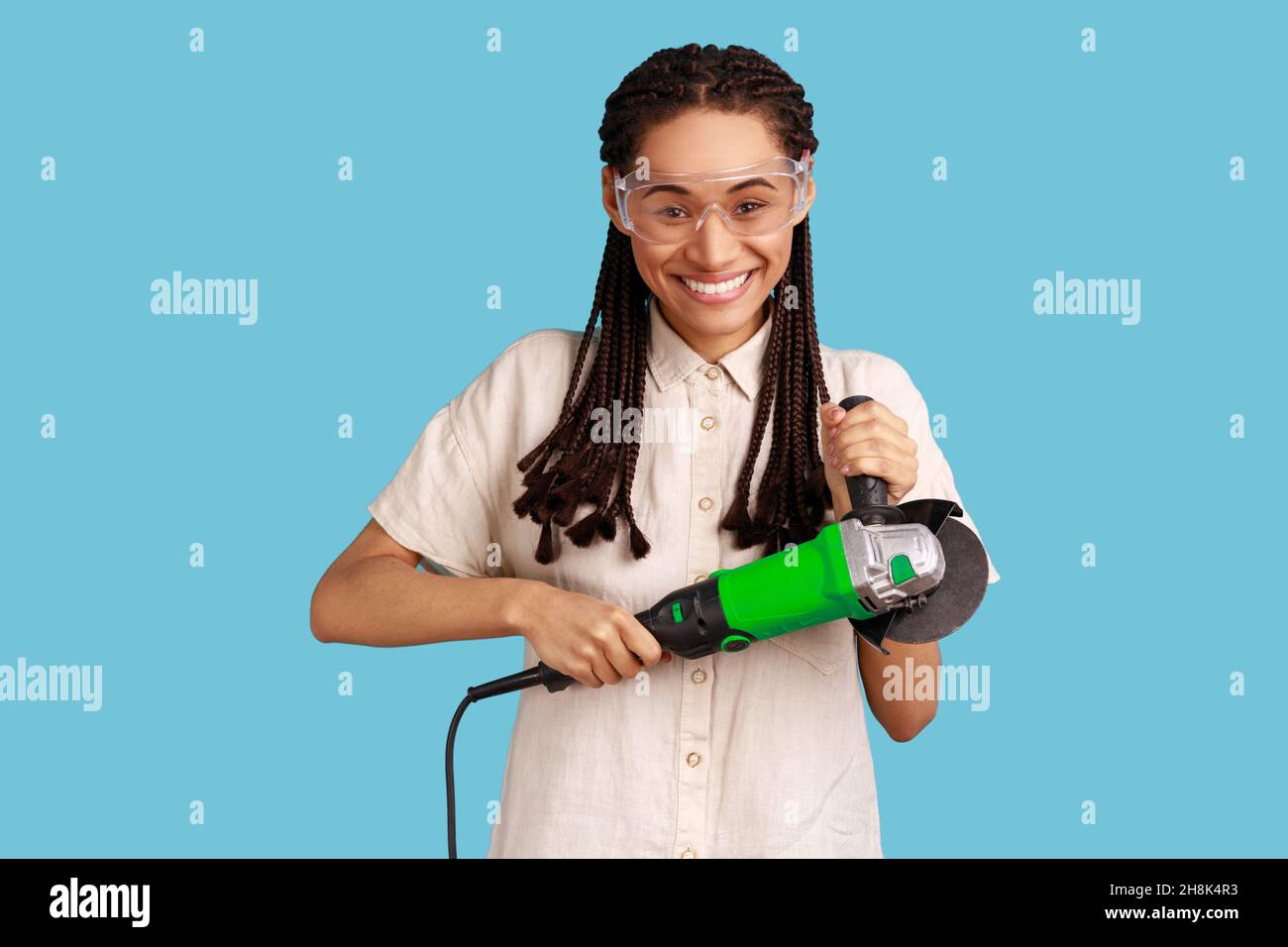 Portrait of smiling woman with dreadlocks holding grinder saw, looking at camera with positive emotions, wearing white shirt and protective glasses. Indoor studio shot isolated on blue background. Stock Photo