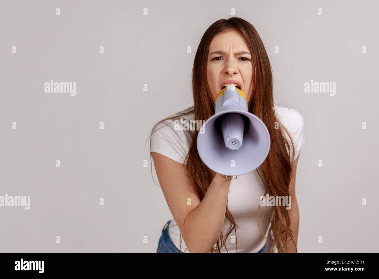 Angry woman holding megaphone near mouth, loudly speaking, screaming, making announcement, paying attention at social problems, wearing white T-shirt. Indoor studio shot isolated on gray background. Stock Photo