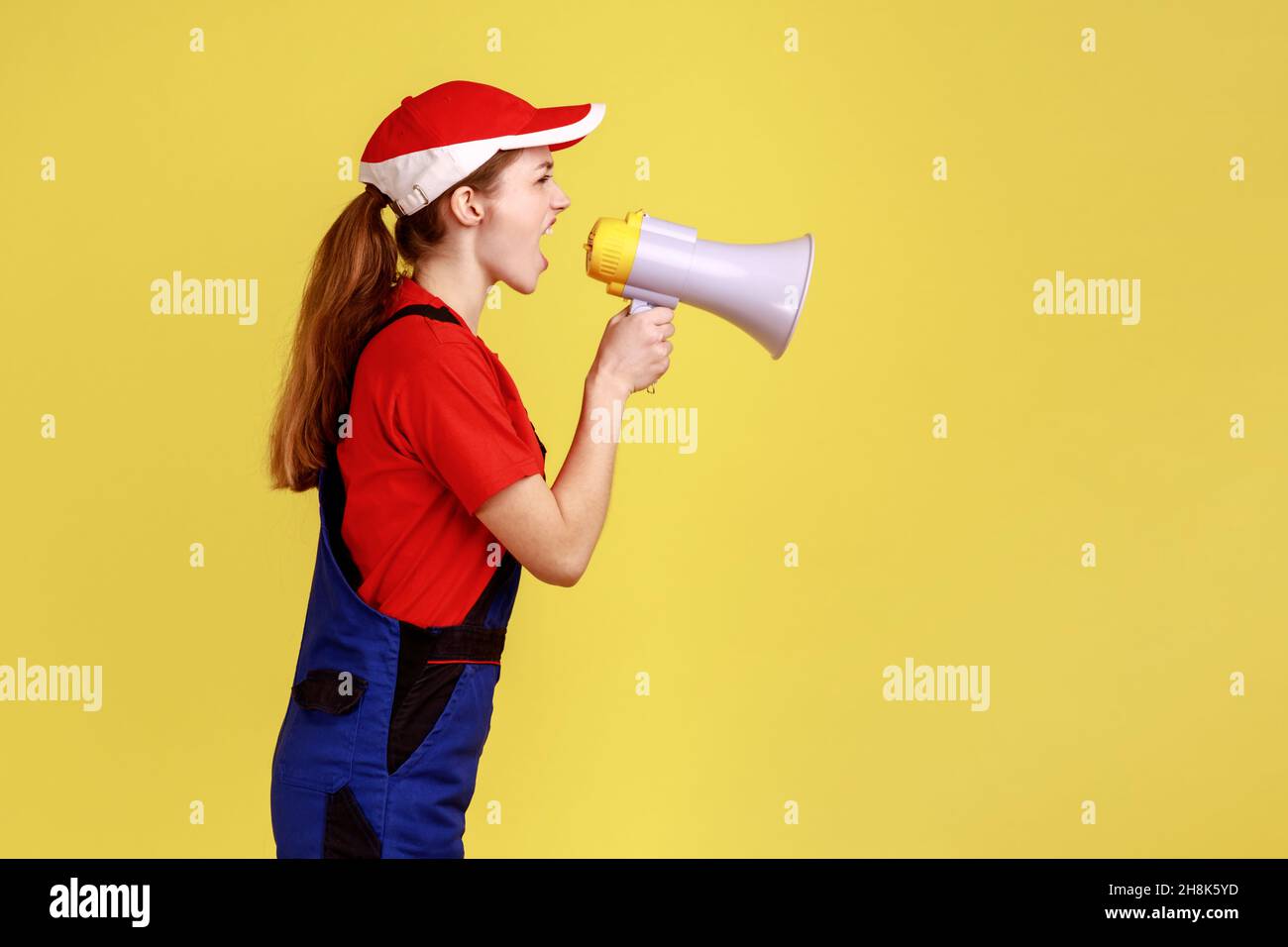 Side view portrait of angry worker woman screaming loud in megaphone, giving command her subordinate builders, wearing work uniform and red cap. Indoor studio shot isolated on yellow background. Stock Photo