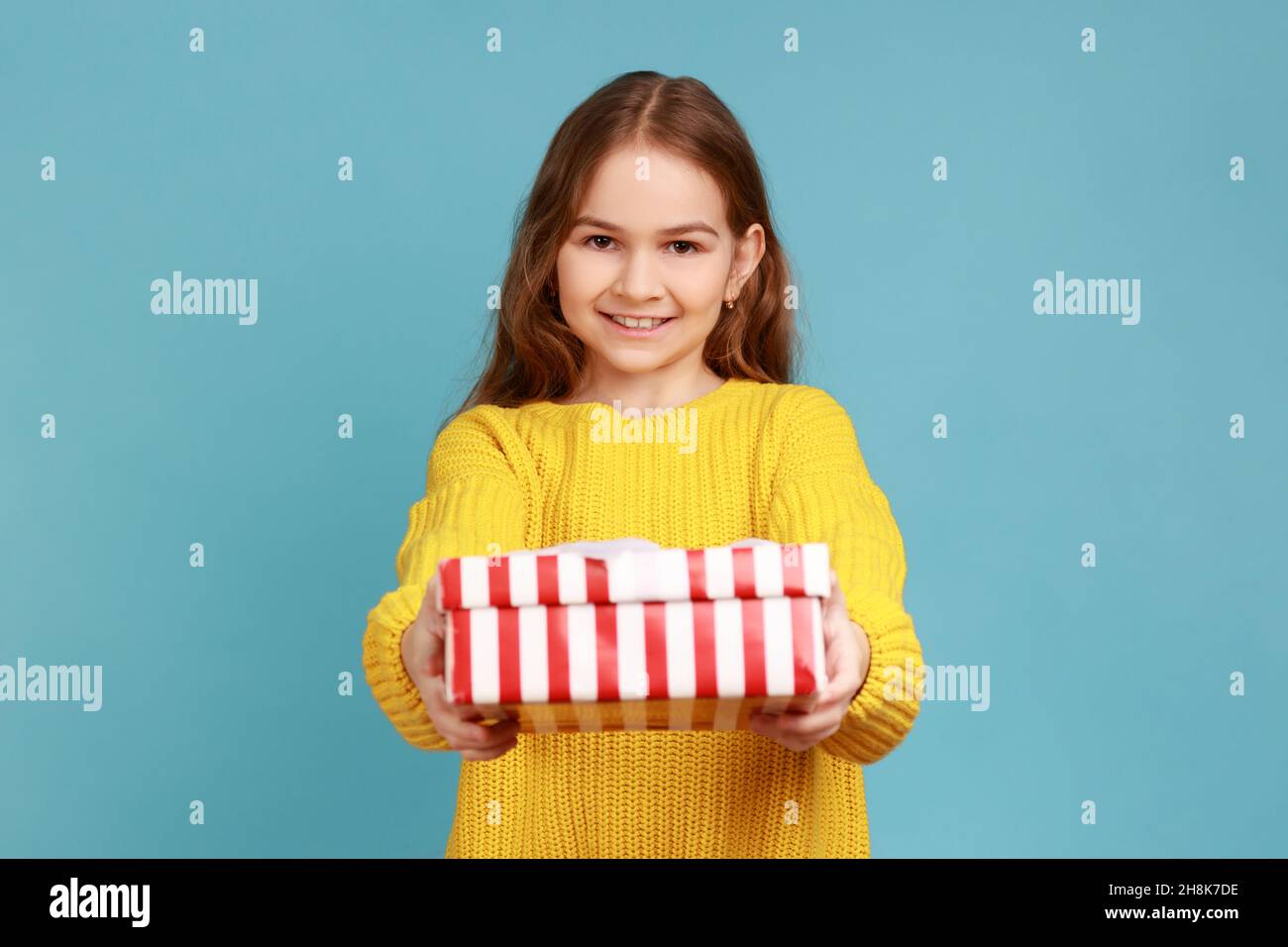 Little girl giving gift box to camera with smile, child greeting on holiday and sharing present, wearing yellow casual style sweater. Indoor studio shot isolated on blue background. Stock Photo