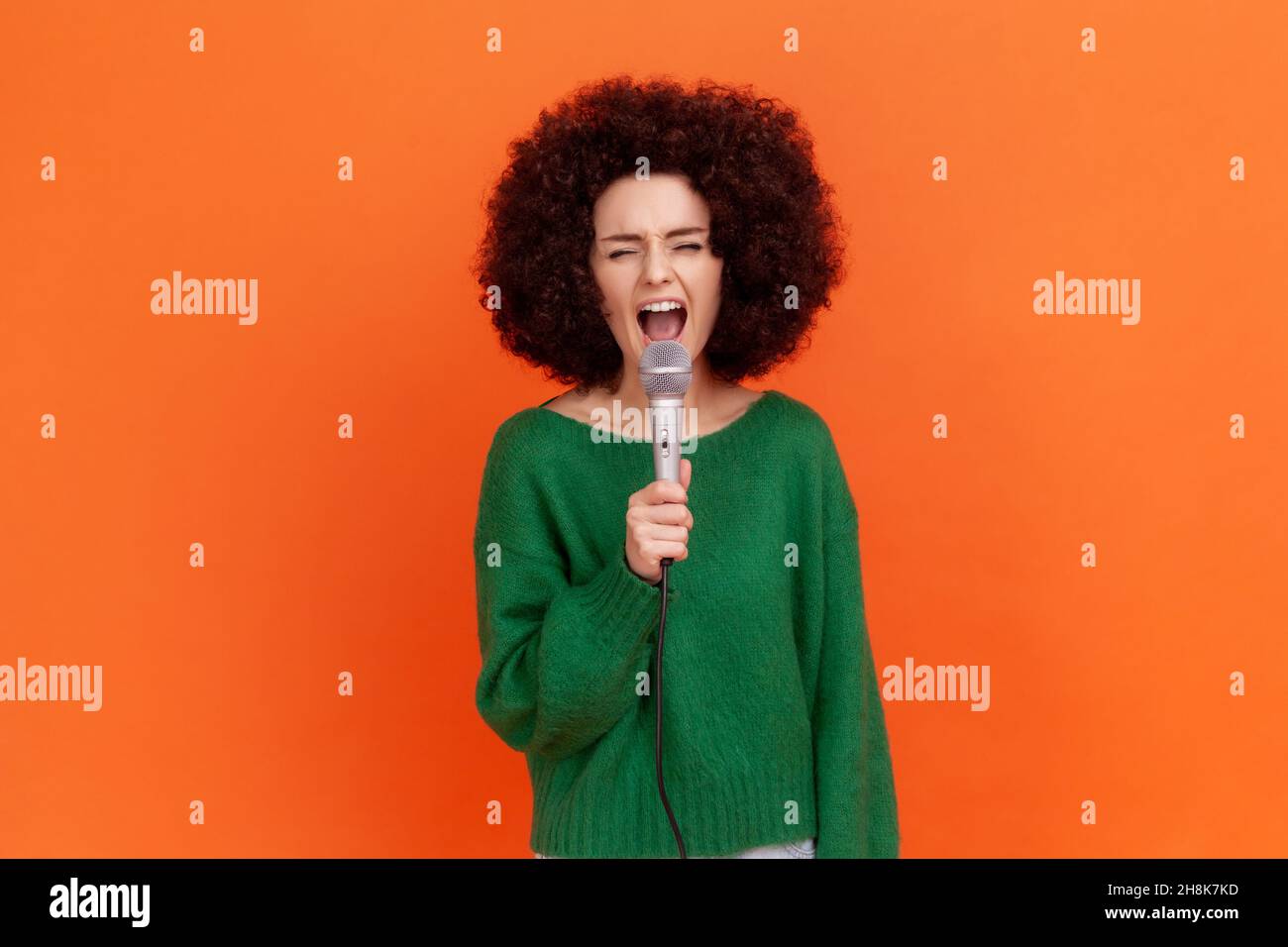 Portrait of attractive woman with Afro hairstyle wearing green casual style sweater singing loud favorite songs with microphone in hands, Indoor studio shot isolated on orange background. Stock Photo