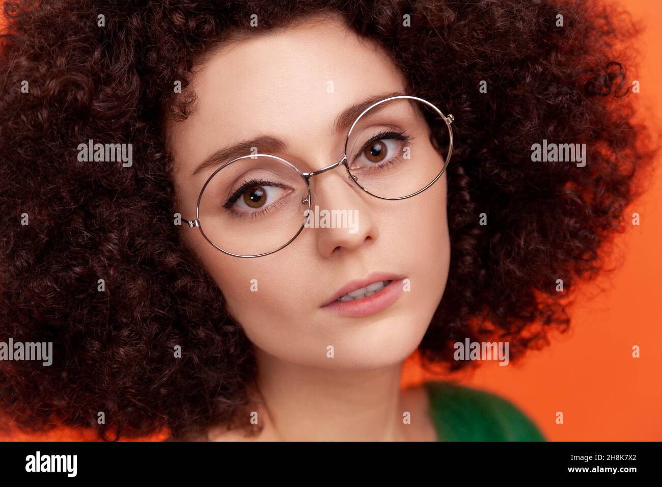 Closeup portrait of attractive woman with Afro hairstyle wearing green casual style sweater and eyeglasses, skin care and beauty concept. Indoor studio shot isolated on orange background. Stock Photo