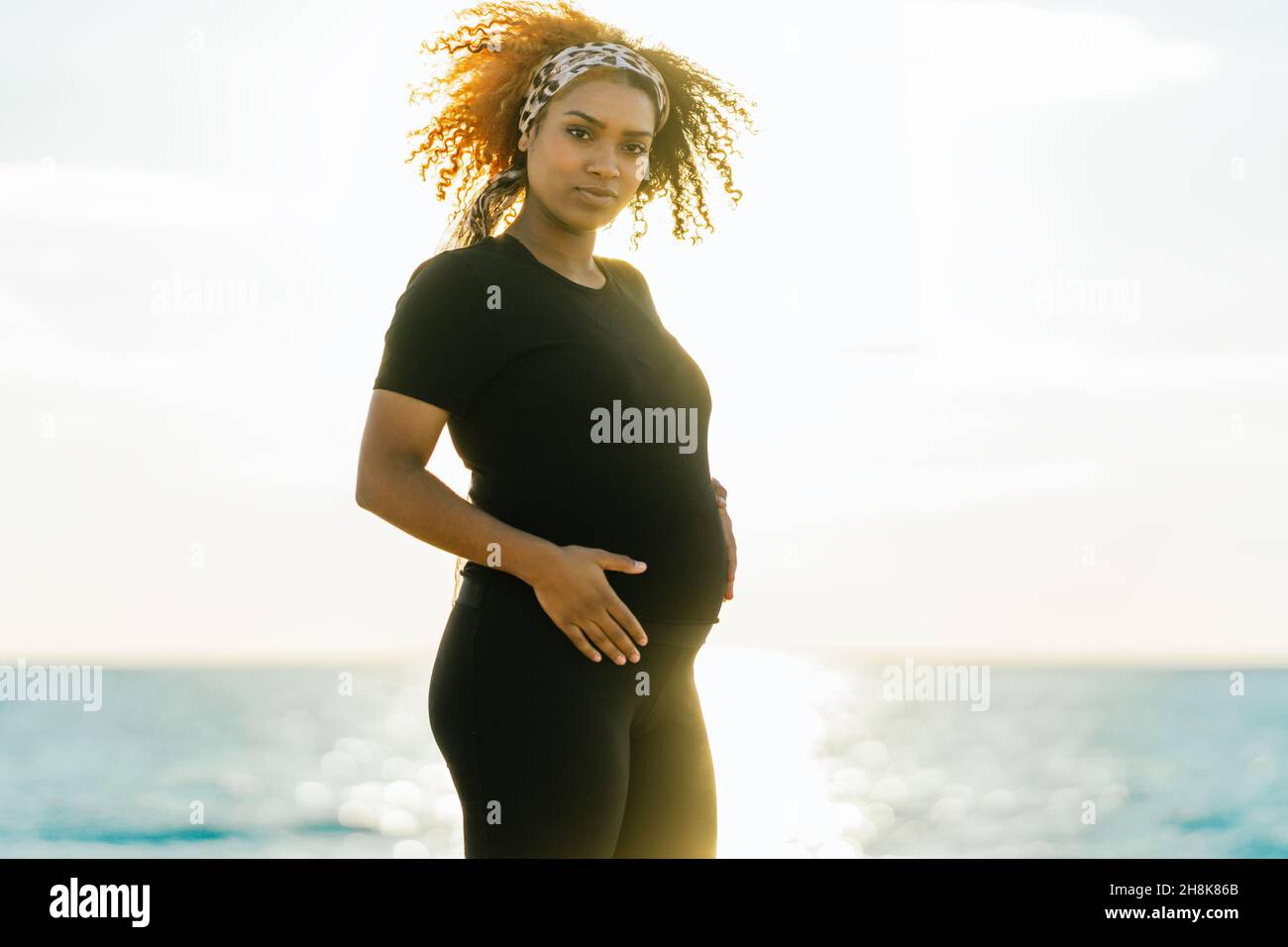 Rays of the sun illuminating a pregnant woman touching her belly on the beach Stock Photo