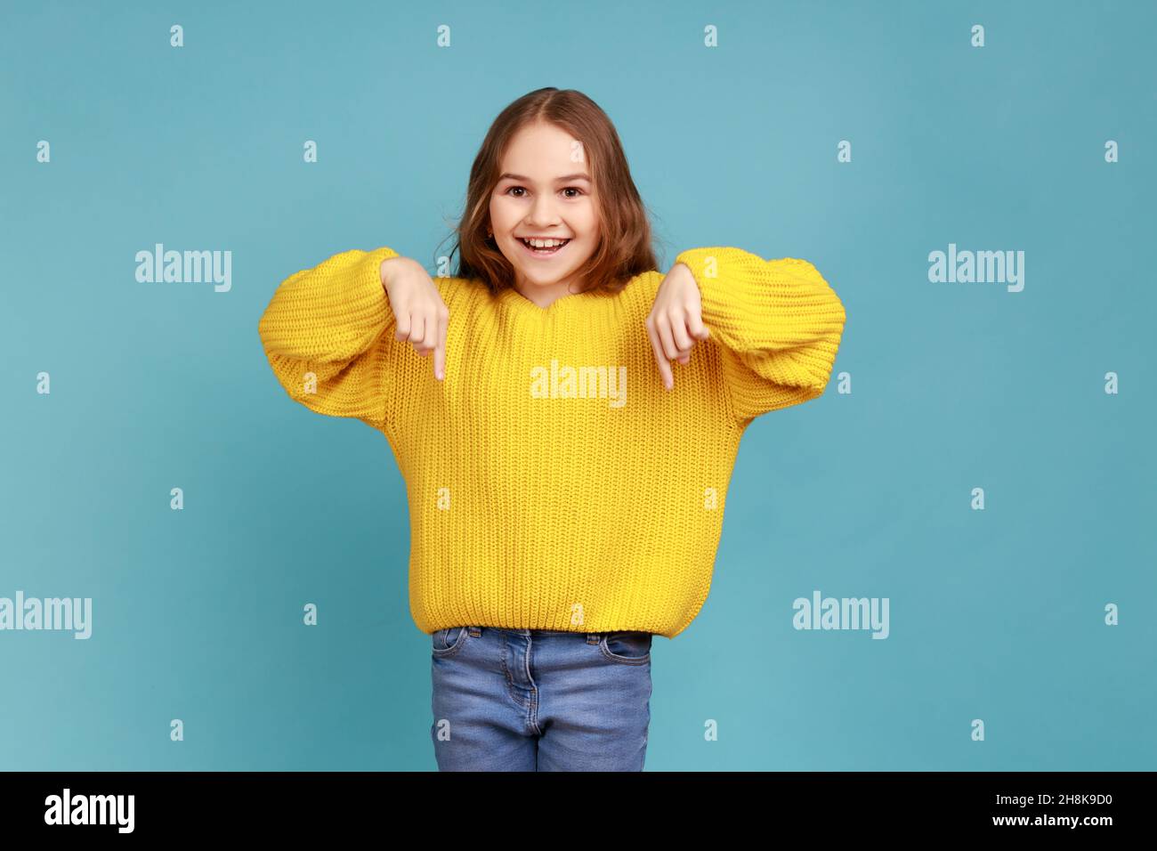 Portrait of cute little girl pointing fingers down, showing place below for commercial information, wearing yellow casual style sweater. Indoor studio shot isolated on blue background. Stock Photo