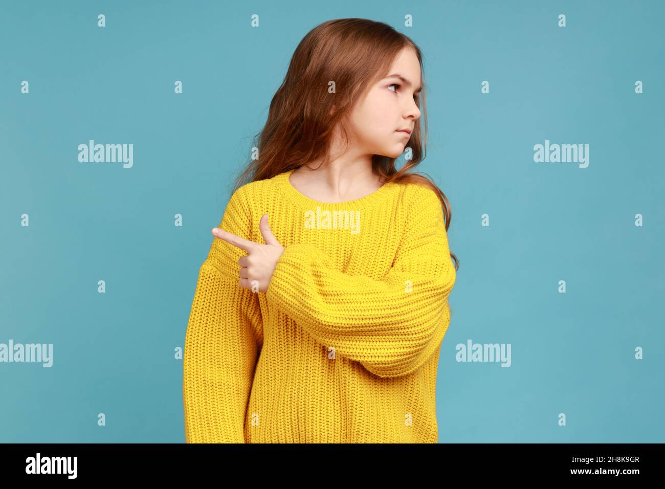 Portrait of little girl pointing way out showing exit, asking to leave her alone, feeling resentful, wearing yellow casual style sweater. Indoor studio shot isolated on blue background. Stock Photo