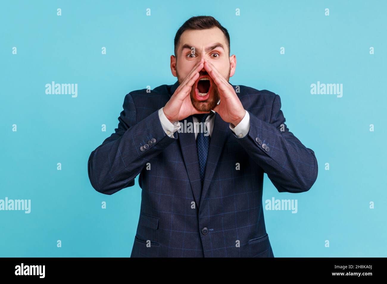 Portrait of hysterical young adult man wearing official style suit loudly yelling in panic, holding hands near his mouth, teenager protest. Indoor studio shot isolated on blue background. Stock Photo