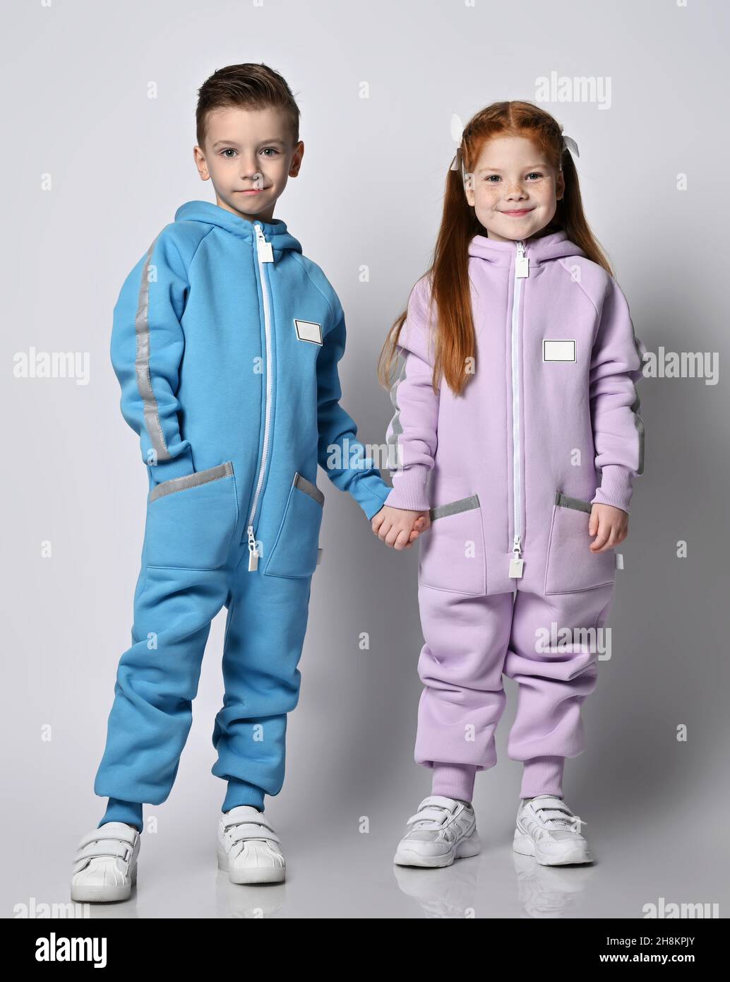 Tow cute smiling kids boy and girl in blue and pink jumpsuits with hoods and pockets stand together holding hands Stock Photo