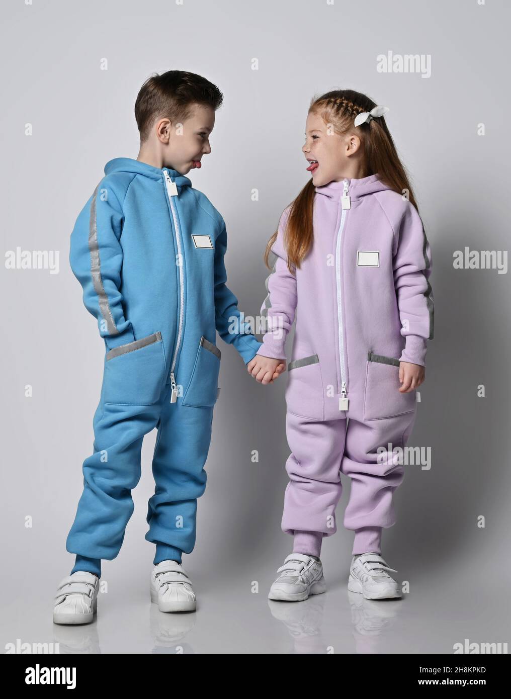 Frolic kids boy and girl, friends in blue and pink jumpsuits stand together holding hands and sticking out thier tongues Stock Photo