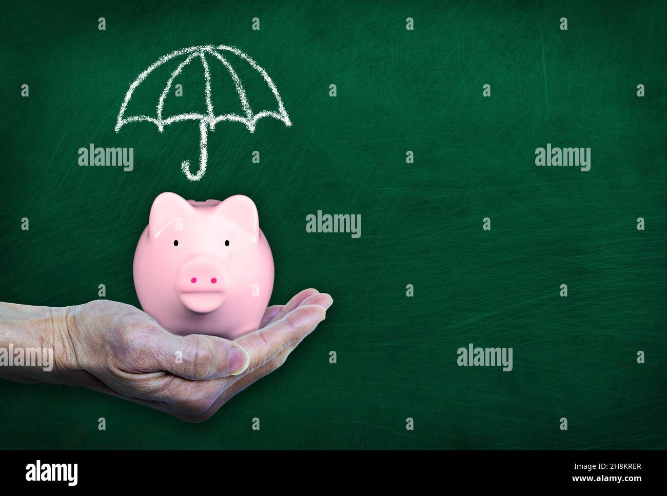 Hand holding pink piggy bank in front of blackboard showing a chalk drawn umbrella. Concept of saving for rainy day, finance insurance, protection, in Stock Photo