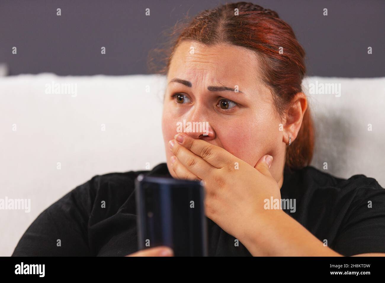 Close up photo of an worried woman in black t-shirt sitting on the white sofa holding the smart phone in her hand and watching bad news on tv covering Stock Photo