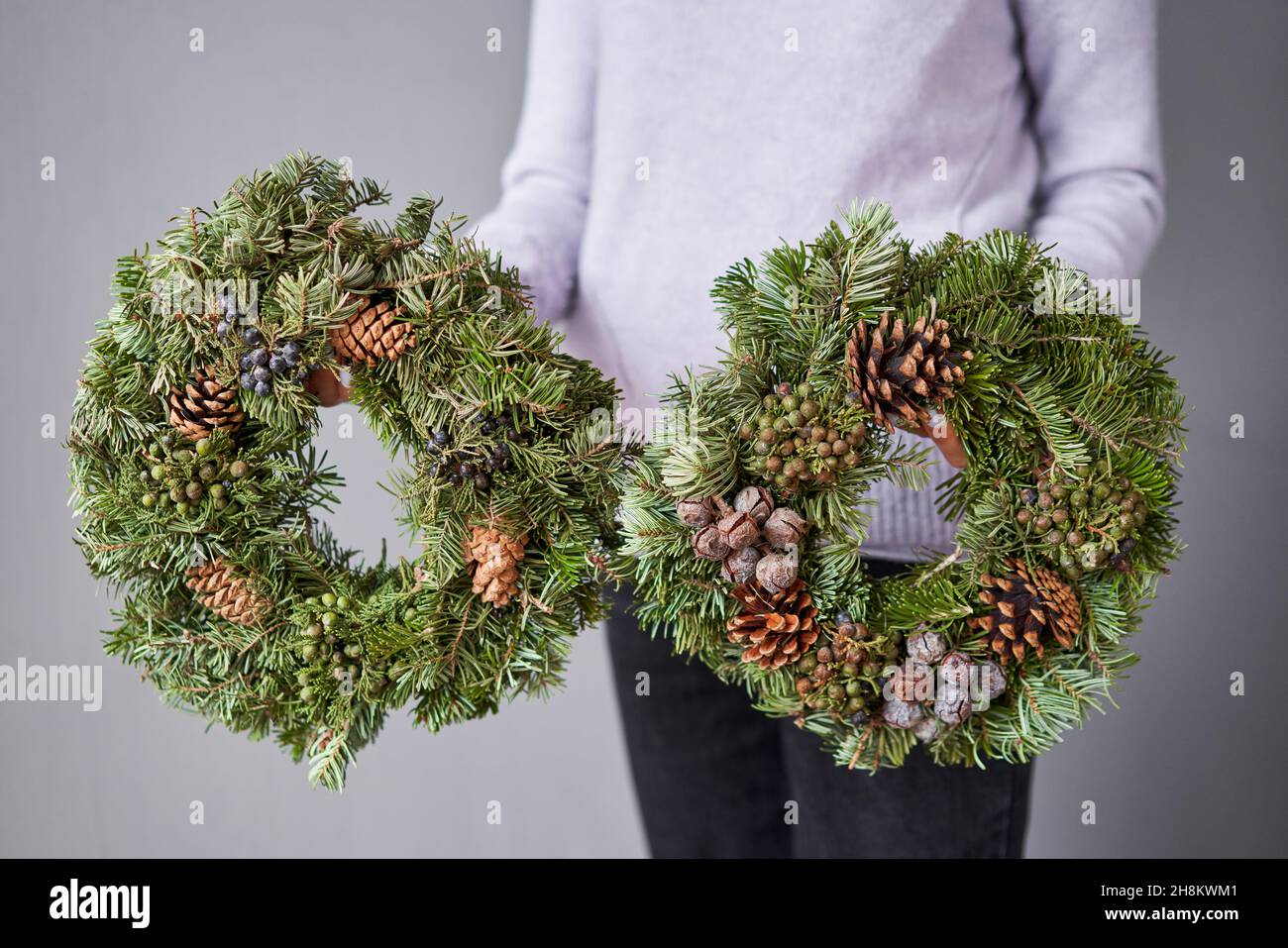 Christmas wreath of fresh spruce in woman hands. Xmas circlet with ornaments, toys and balls. Christmas mood. Gray wall on background Stock Photo