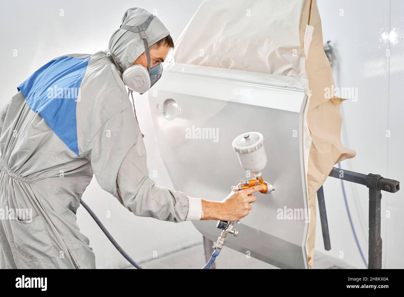 Painting the rear door of the car. Car painter in special painting chamber, wearing costume and protective gear. Car service station. Restoring a car Stock Photo