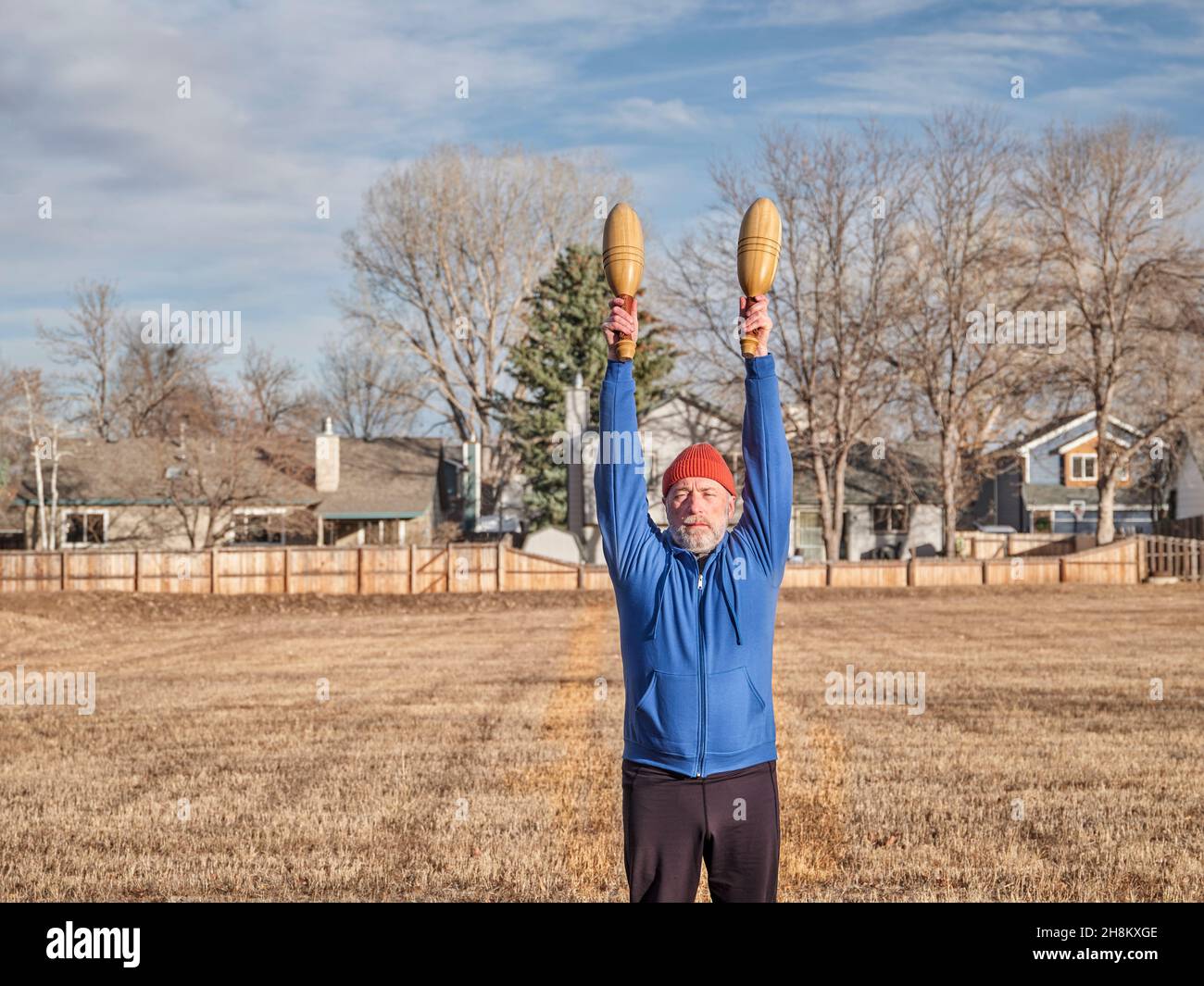 Senior Athletic Male Is Exercising With Wooden Indian Clubs In A Senior athletic male is exercising with wooden indian clubs in a