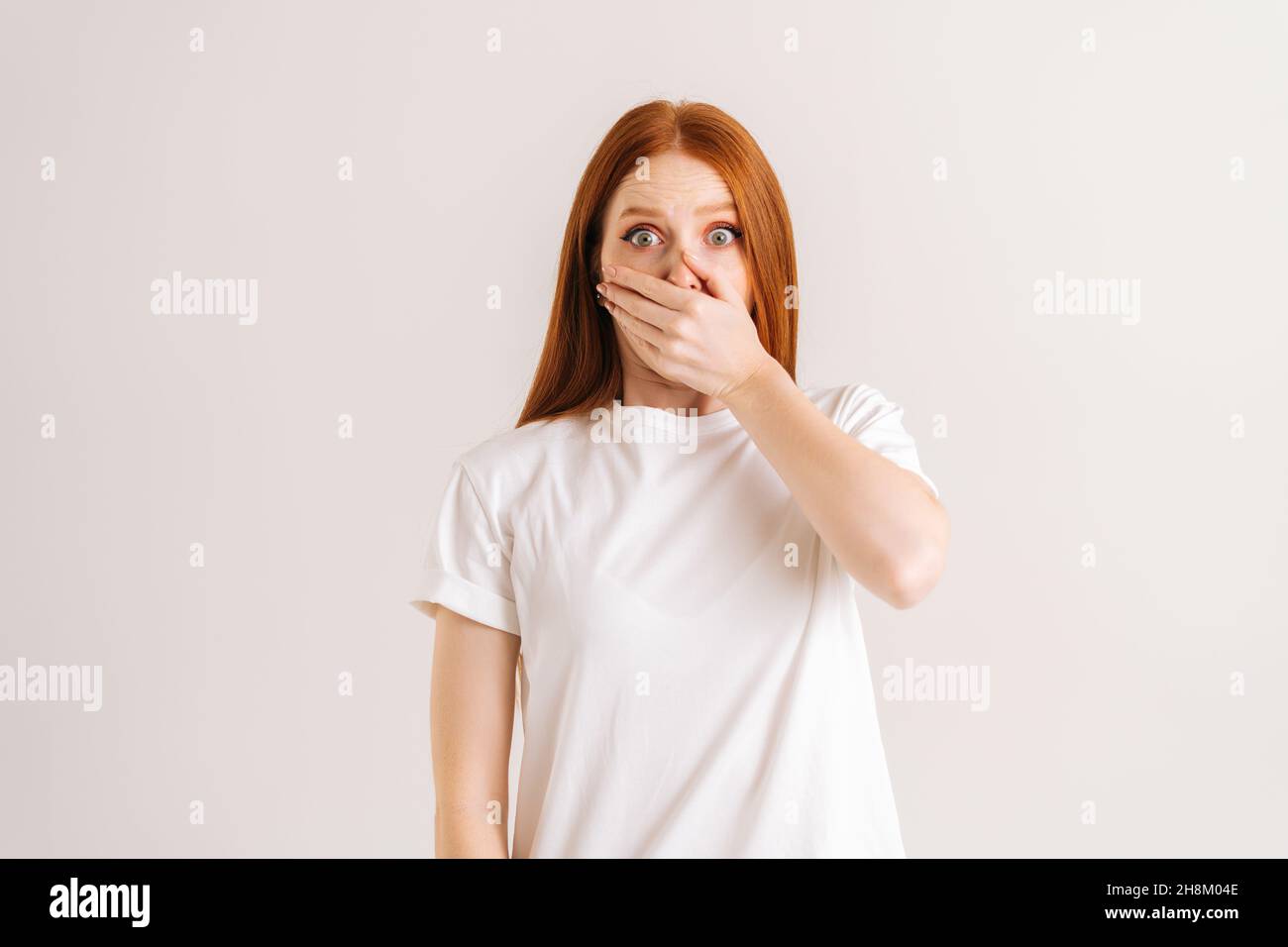 Studio portrait of astonished shocked young woman covering mouth with pressed palms popping eyes at camera from surprise and worry Stock Photo