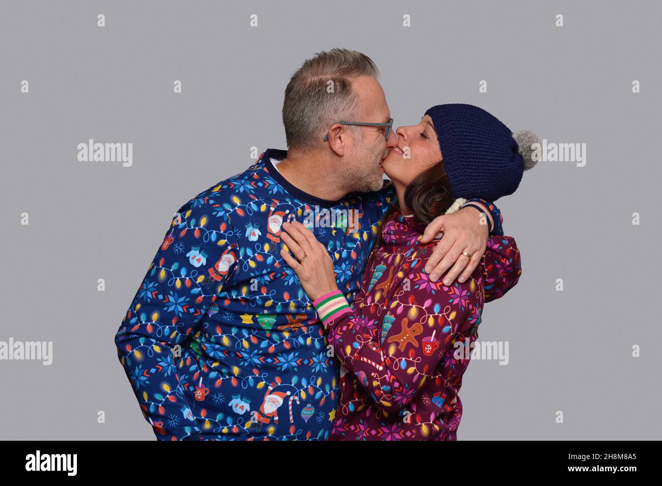 Happy middle-aged couple kissing as they celebrate Christmas together dressed in colorful Xmas themed clothing posing in front of a grey wall Stock Photo