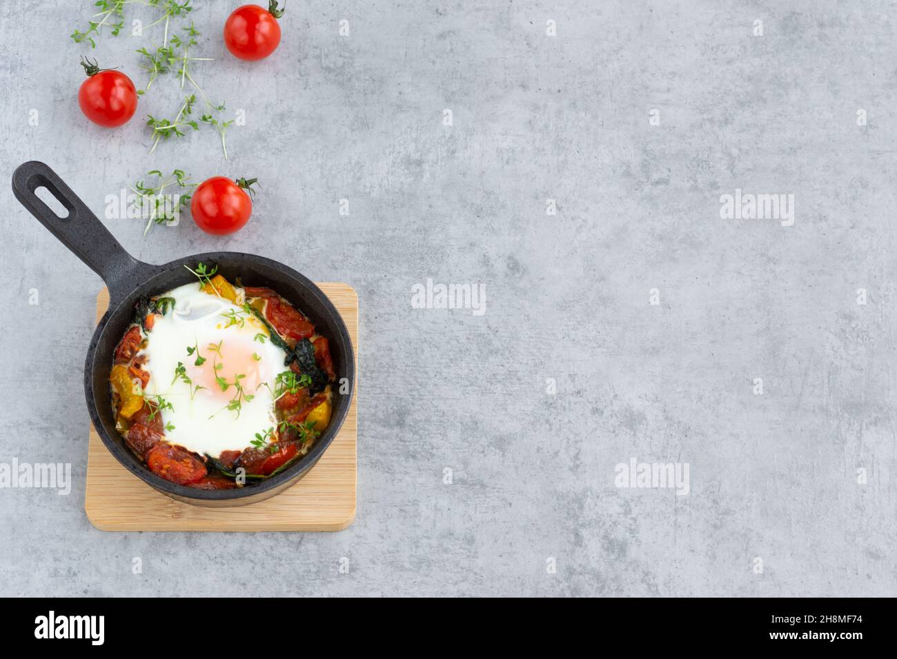 Shakshuka in a skillet on grey background with copy space. Baked egg with vegetables: tomatoes, bell pepper, spinach and garden cress. Stock Photo