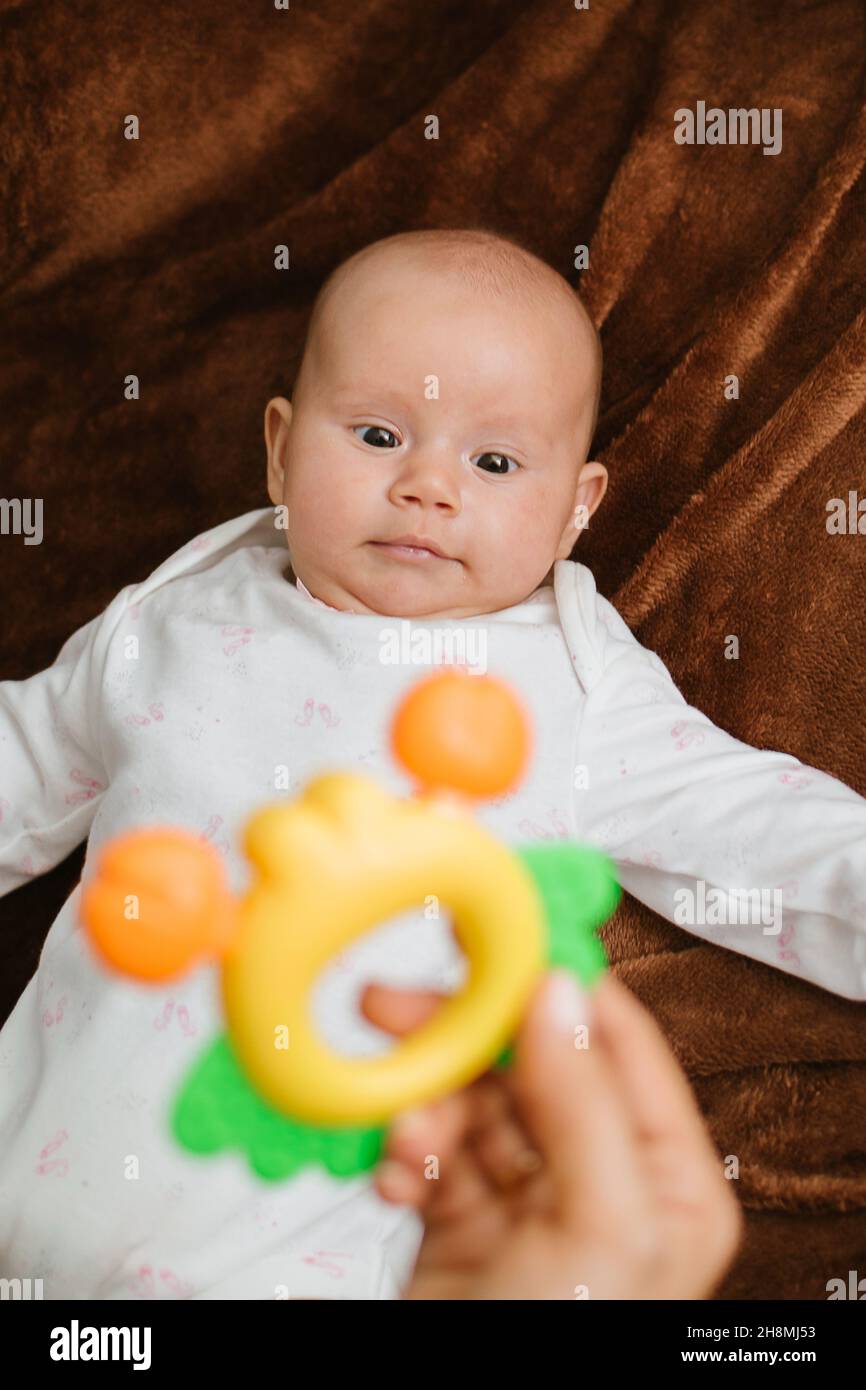 First Rattle Toy for Baby. Rattles and Teethers, Teething Toys. Cute newborn baby girl looking at mothers hand with bright rattle. Selective focus Stock Photo