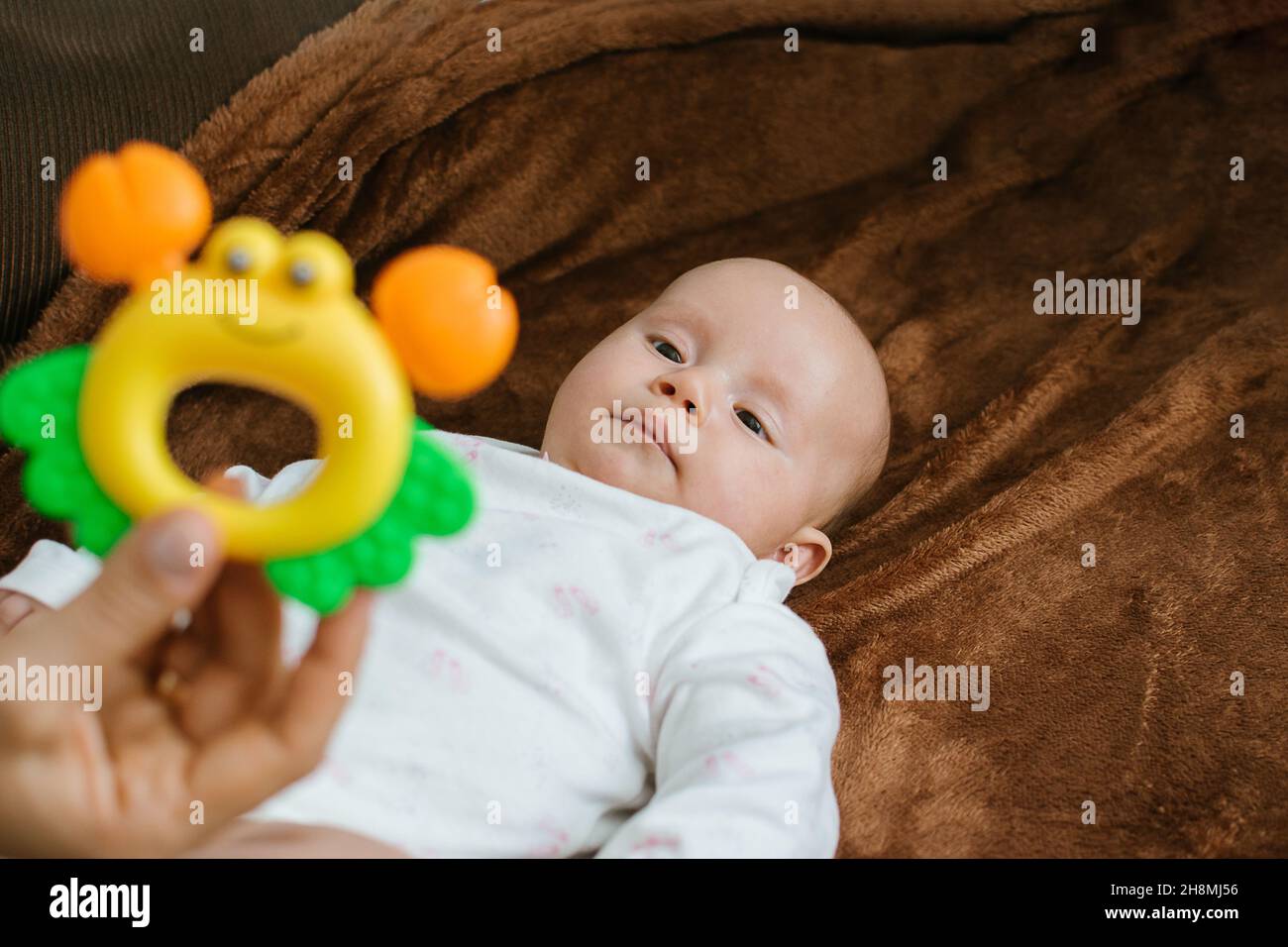 First Rattle Toy for Baby. Rattles and Teethers, Teething Toys. Cute newborn baby girl looking at mothers hand with bright rattle. Selective focus Stock Photo