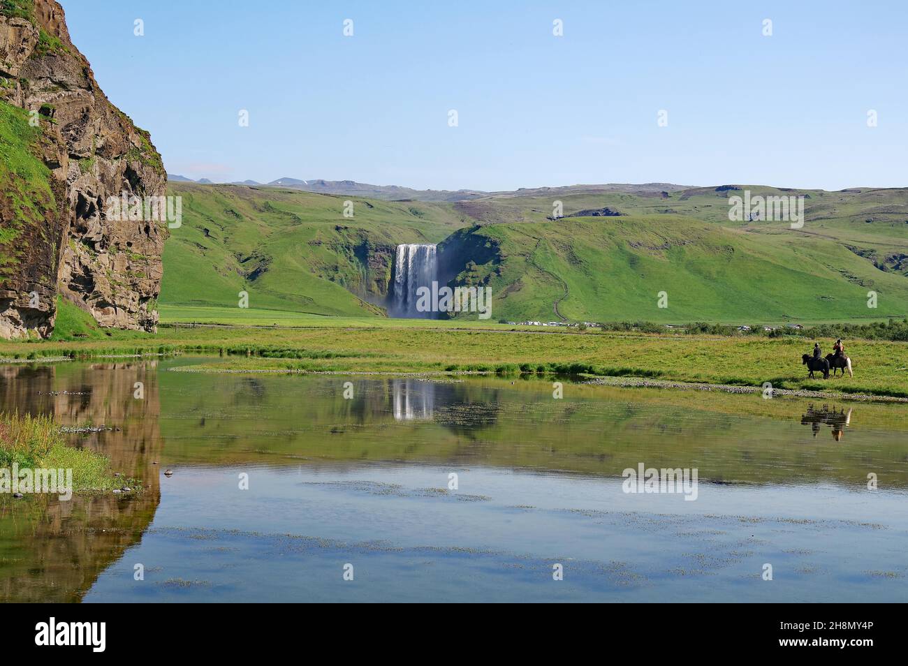 Water masses plummeting vertically into the depths, reflection in the water, green landscape, two riders on Icelandic horses, Skogafoss, Iceland Stock Photo