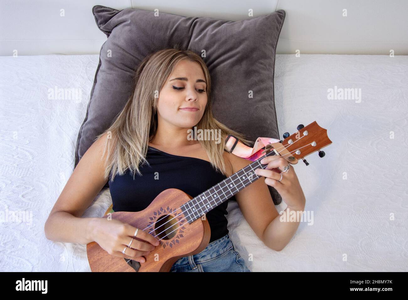 Woman playing the ukulele sitting on the bed in her room at home Stock