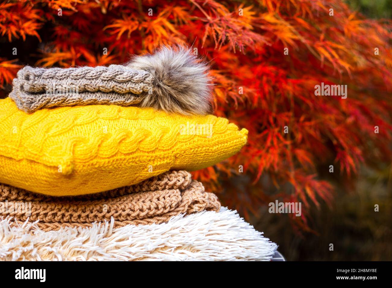 Stack of knitted warm clothing and wool pillow. Autumn leaf background. Knit hat with pom pom and sweater Stock Photo