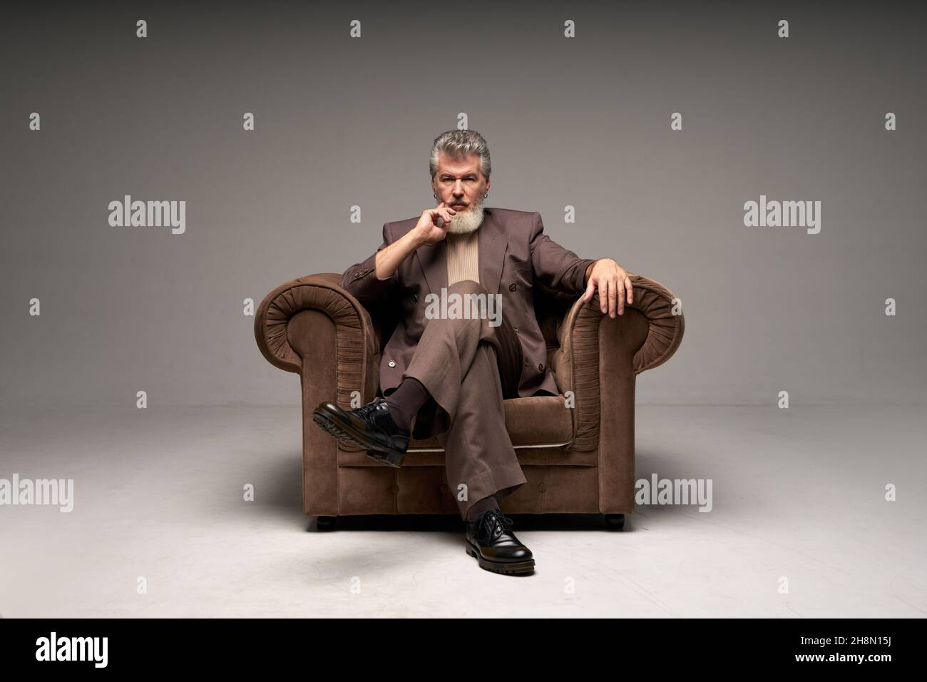 Full length shot of pensive mature businessman with beard wearing elegant suit looking at camera, sitting in armchair while posing isolated over light Stock Photo