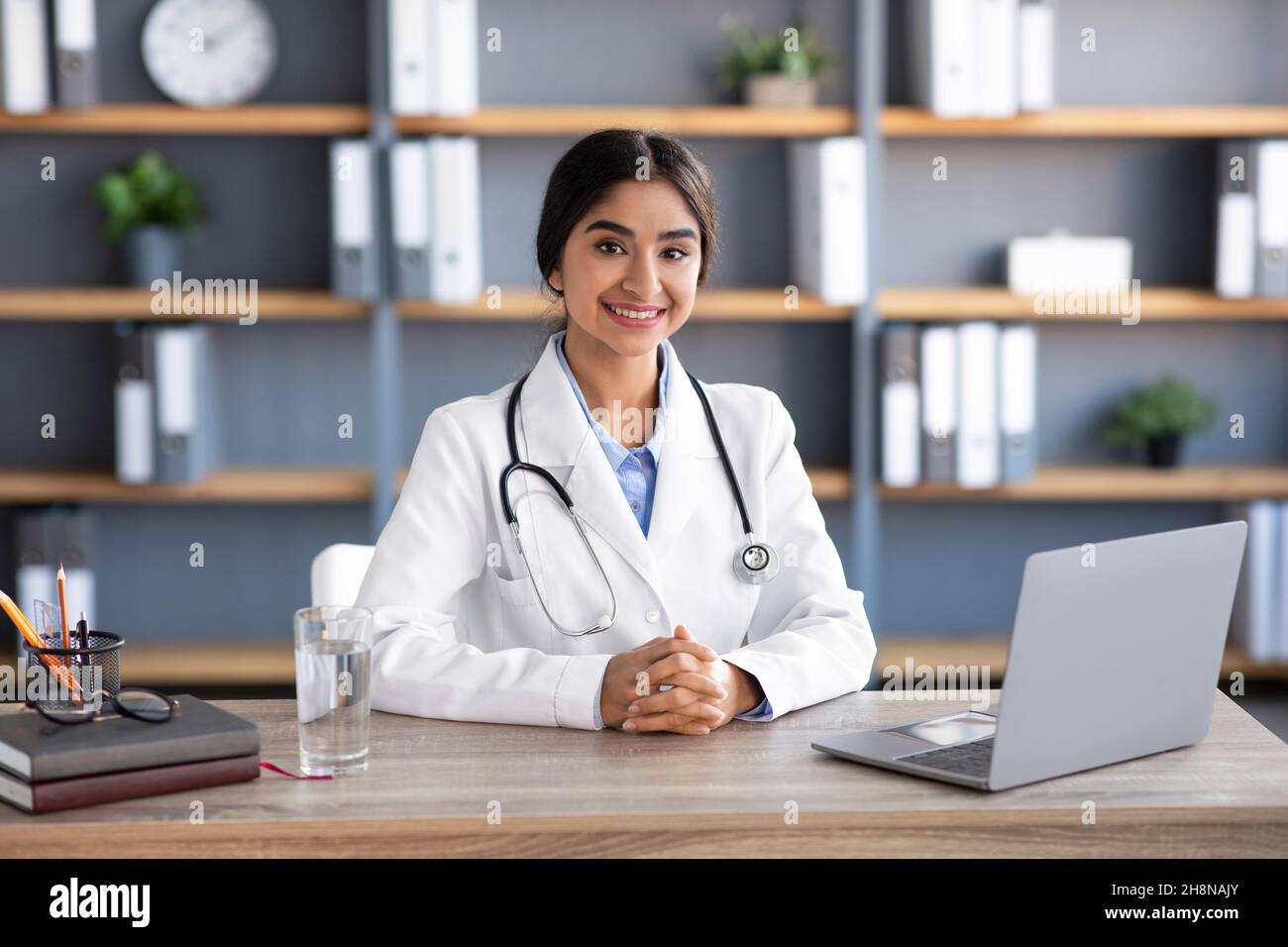Smiling friendly young indian female doctor in white coat at workplace ...
