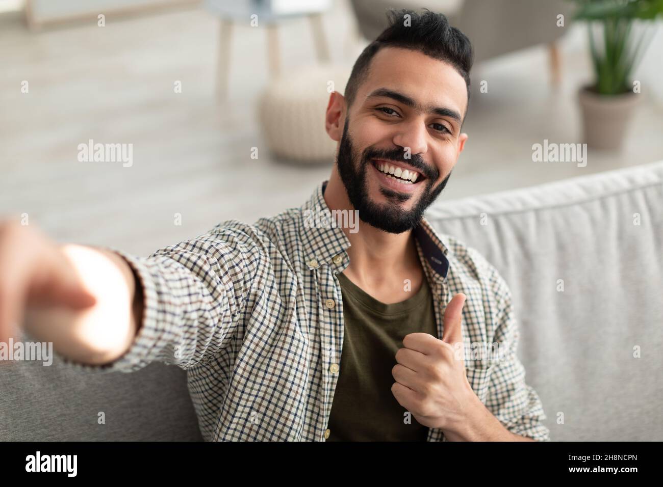 Happy young Arab guy taking selfie and showing thumb up gesture, smiling at mobile phone camera, sitting on sofa at home Stock Photo