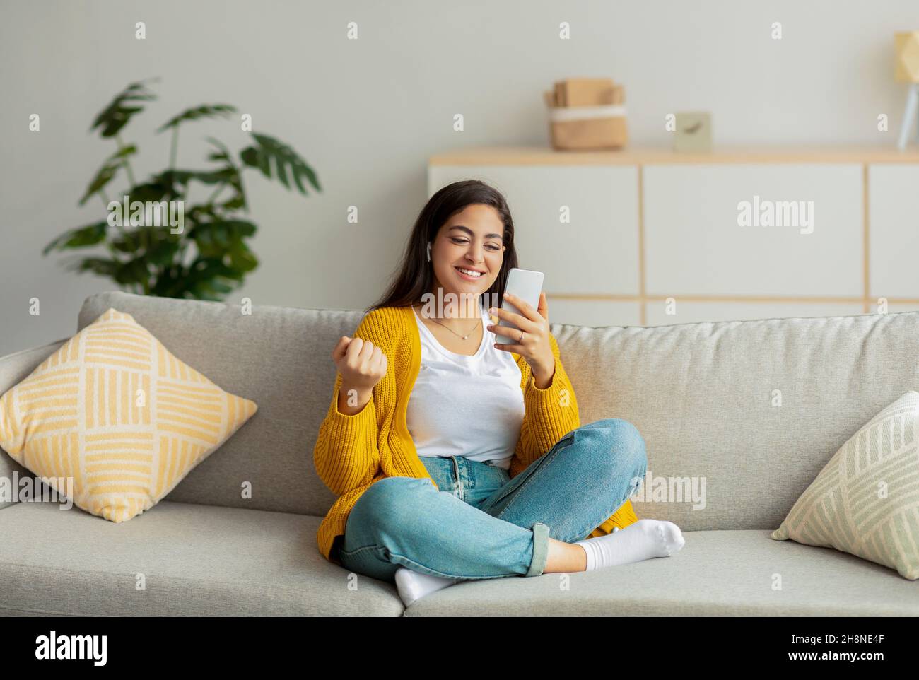 Yes. Excited arab woman using smartphone gadget, shaking clenched fist, celebrating victory, wearing wireless earbuds Stock Photo