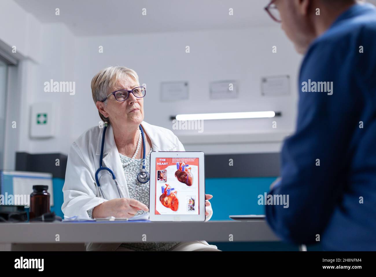 Specialist talking about cardiology diagnosis with image of heart organ on tablet to man. General practitioner showing illustration on cardiovascular system and anatomical structure to patient. Stock Photo